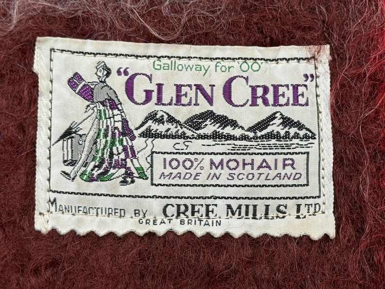 May include: A rectangular white label with the text "Galloway for Glen Cree" in purple and green. The label features an illustration of a person in a kilt and mountains. The label reads "100% Mohair Made in Scotland" and "Manufactured by Cree Mills Ltd Great Britain."
