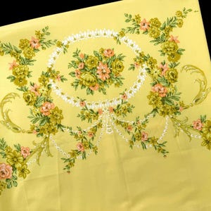 May include: A pale yellow fabric featuring a central floral arrangement surrounded by a white decorative oval frame. The design includes cascading floral garlands in shades of green, olive, and soft pink, accented with white beaded swags.