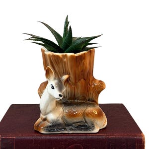 May include: A ceramic planter shaped like a tree stump with a resting deer figure at the base. The deer is white, brown, and gray, while the stump is textured brown. A small green succulent plant is potted inside the hollow top of the stump.