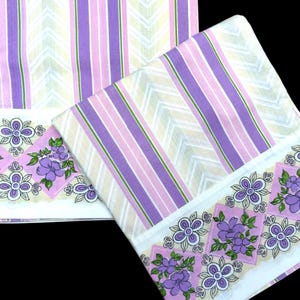 May include: Two white cotton pillowcases with purple and pink stripes and a floral border. The floral border features purple flowers and green leaves.
