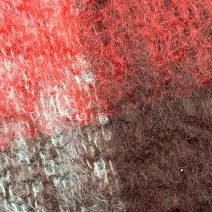 May include: Close-up of a textured fabric with a mix of red, brown, and gray hues. The fibers are densely packed, creating a soft, fuzzy appearance. The color variations create a visually interesting pattern.