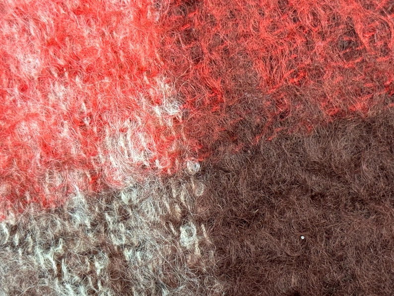 May include: Close-up of a textured fabric with a mix of red, brown, and gray hues. The fibers are densely packed, creating a soft, fuzzy appearance. The color variations create a visually interesting pattern.