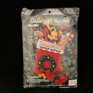 May include: A red felt stocking with a wreath and bells, filled with felt toys, including a gingerbread man, a doll, and a rocking horse. The stocking is labeled "Heather" and is part of the Gallery of Stitches by Bucilla Christmas Surprises collection.