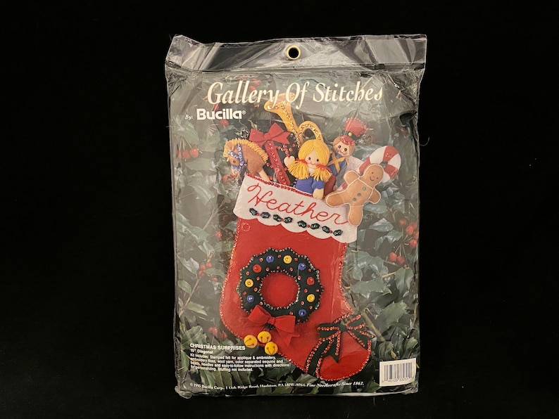 May include: A red felt stocking with a wreath and bells, filled with felt toys, including a gingerbread man, a doll, and a rocking horse. The stocking is labeled "Heather" and is part of the Gallery of Stitches by Bucilla Christmas Surprises collection.