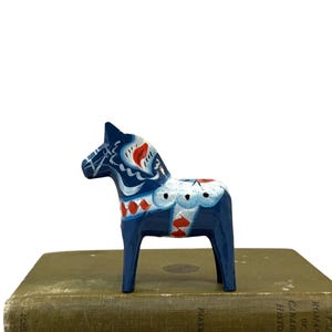 May include: A dark blue wooden horse figurine featuring hand-painted white and red floral and geometric patterns on its mane, neck, and back. The horse stands in a profile position with a smooth, glossy finish and detailed decorative brushwork.