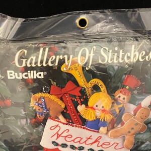 May include: A Bucilla Gallery of Stitches Christmas stocking kit featuring a red and green stocking with a name "Heather" embroidered on the front. The stocking is decorated with a variety of felt ornaments including a gingerbread man, a toy soldier, a rocking horse, a girl doll, and a trumpet.