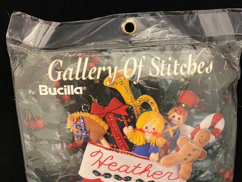 May include: A Bucilla Gallery of Stitches Christmas stocking kit featuring a red and green stocking with a name "Heather" embroidered on the front. The stocking is decorated with a variety of felt ornaments including a gingerbread man, a toy soldier, a rocking horse, a girl doll, and a trumpet.