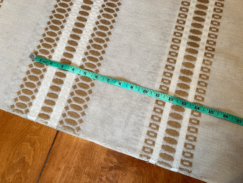 May include: A beige fabric with a brown geometric pattern. A measuring tape is laid across the fabric, showing a measurement of 17 inches.