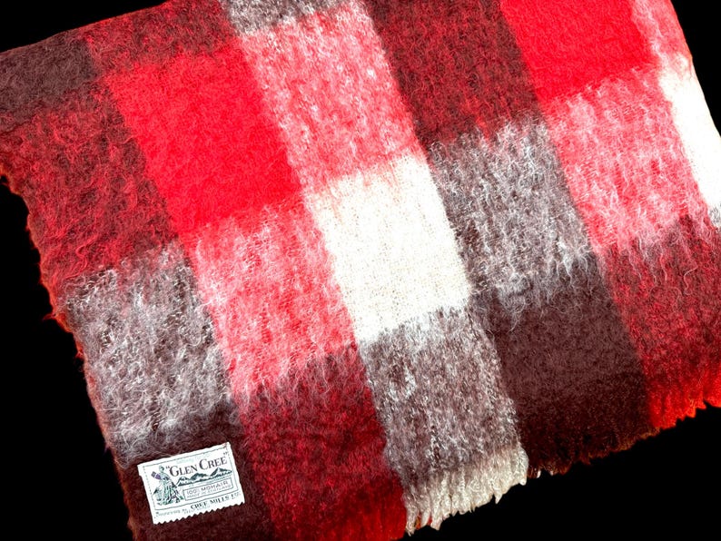 May include: A close-up of a red, white, and brown plaid blanket. The blanket has a soft, fuzzy texture and a rectangular shape. A small tag with text is visible in the corner.