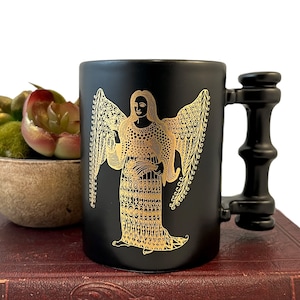 K&ouml;nnte beinhalten: Schwarze Keramiktasse mit einem goldenen Engelmotiv. Die Tasse hat einen schwarzen Henkel mit einem einzigartigen Design.