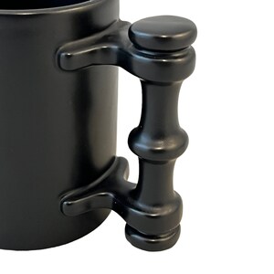 K&ouml;nnte beinhalten: Schwarze Keramiktasse mit einem goldenen Design eines stilisierten Elefanten. Die Tasse hat einen einzigartigen, schwarzen Griff im Bambus-Stil.