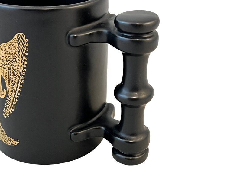 K&ouml;nnte beinhalten: Schwarze Keramiktasse mit einem goldenen Design eines stilisierten Elefanten. Die Tasse hat einen einzigartigen, schwarzen Griff im Bambus-Stil.