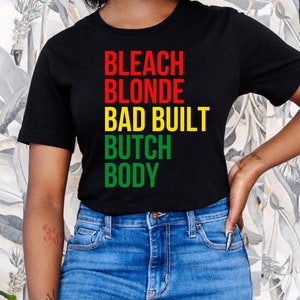 Könnte beinhalten: Ein schwarzes T-Shirt mit dem Text "BLEACH BLONDE BAD BUILT BUTCH BODY" in Rot, Gelb und Grün.