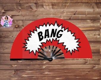 May include: A red hand fan with the word "BANG" in bold black letters, set against a white burst design. The fan's frame is black, and the background is a wooden surface.