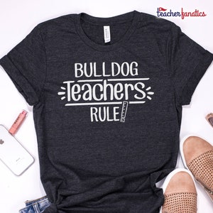 Pu&ograve; includere: Maglietta grigio scuro con testo bianco che recita "Bulldog Teachers Rule" con un grafico di righello.