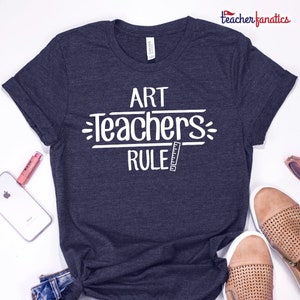 Könnte beinhalten: Ein dunkelblaues T-Shirt mit weißem Text, der "Art Teachers Rule" lautet, mit einer Linealgrafik.
