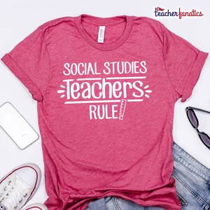 May include: A red t-shirt with the text "SOCIAL STUDIES TEACHERS RULE" in white. The shirt has rolled-up sleeves and is displayed with a smartphone, sunglasses, and a pair of white sneakers.