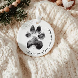 May include: A round, white ceramic ornament featuring a black paw print and a white ribbon. The text "Winnie, you left paw prints on our hearts" is written in a handwritten style. A pet memorial ornament.