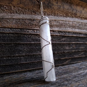 May include: A white selenite crystal wrapped in silver wire, hanging from a silver chain. The crystal is smooth and polished, with a natural, striated texture. It is hanging against a rustic wooden background.
