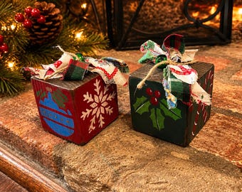 May include: Two square wooden blocks painted with holiday motifs. The red block features a blue ornament and a white snowflake. The dark green block displays holly leaves with red berries and candy canes. Both are topped with fabric bows.