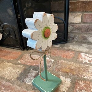 May include: A white wooden daisy with a green center and a brown button. The daisy is attached to a green wooden base with a brown twine bow.