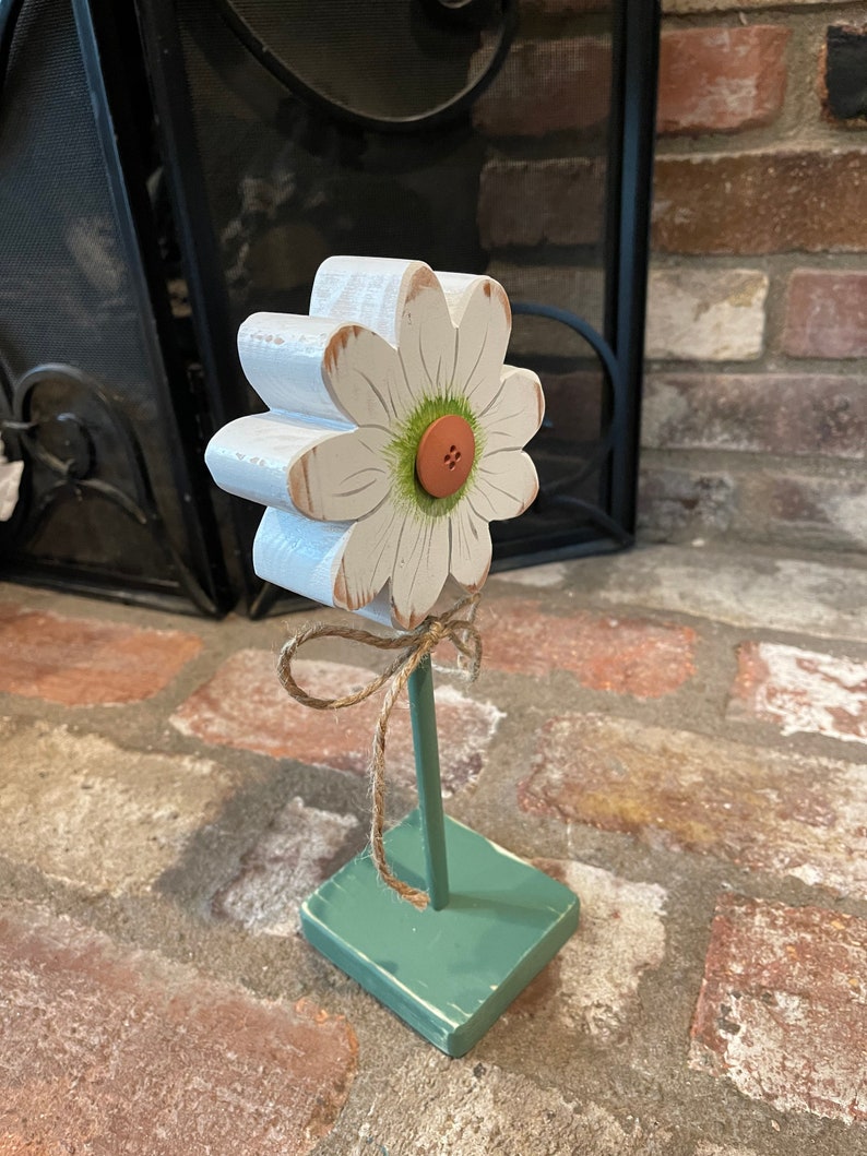 May include: A white wooden daisy with a green center and a brown button. The daisy is attached to a green wooden base with a brown twine bow.