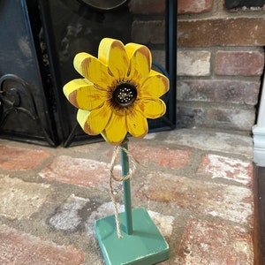 May include: A wooden sunflower with yellow petals and a brown center on a green painted wooden stand. The sunflower is tied to the stand with twine.