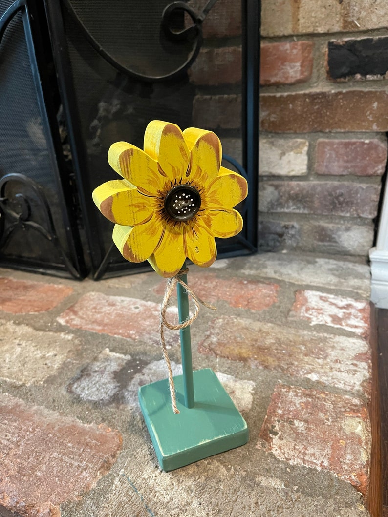 May include: A wooden sunflower with yellow petals and a brown center on a green painted wooden stand. The sunflower is tied to the stand with twine.