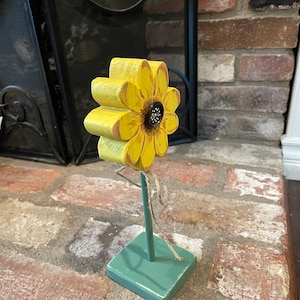 May include: A wooden sunflower with yellow petals and a brown center, standing on a green painted wooden base. The sunflower is attached to the base with a brown twine.