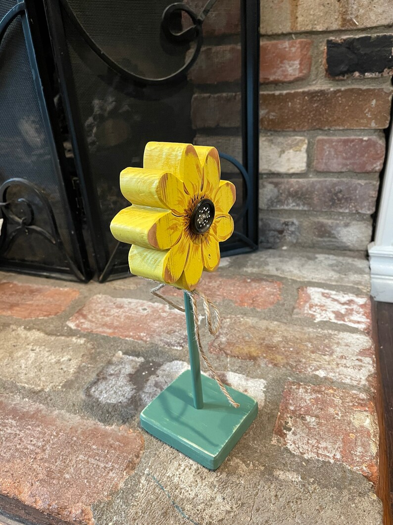 May include: A wooden sunflower with yellow petals and a brown center, standing on a green painted wooden base. The sunflower is attached to the base with a brown twine.