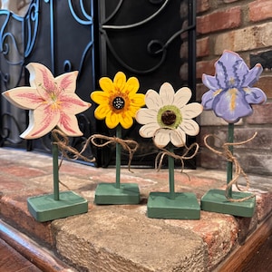 May include: Four wooden flower figurines with painted details. The flowers are a pink lily, a yellow sunflower, a white daisy, and a purple iris. Each flower is on a green painted stick with a green square base.