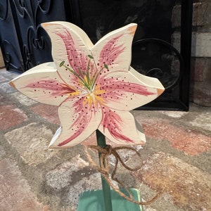May include: A wooden lily flower with pink and white petals and a yellow center. The flower is painted with details and is attached to a green wooden stick. The stick is tied with twine.