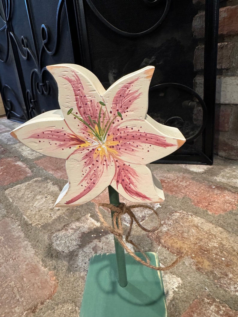 May include: A wooden lily flower with pink and white petals and a yellow center. The flower is painted with details and is attached to a green wooden stick. The stick is tied with twine.