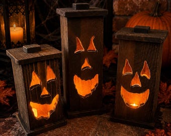 Puede incluir: Tres linternas rectangulares de madera marrón oscuro con caras de calabaza talladas. Cada linterna tiene una parte superior y una base planas, con una luz naranja cálida que brilla desde el interior a través de los ojos, narices y bocas.