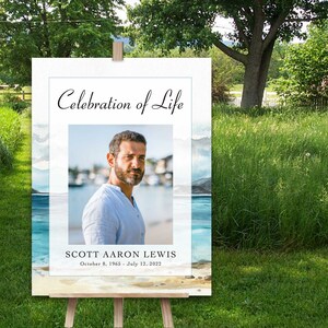 May include: A white sign with a watercolour background of a beach scene. The sign says "Celebration of Life" and features a photo of a man with a beard. The text below the photo reads "Scott Aaron Lewis, October 8, 1965 - July 12, 2022".