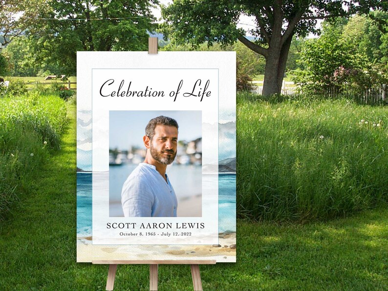 May include: A white sign with a watercolour background of a beach scene. The sign says "Celebration of Life" and features a photo of a man with a beard. The text below the photo reads "Scott Aaron Lewis, October 8, 1965 - July 12, 2022".