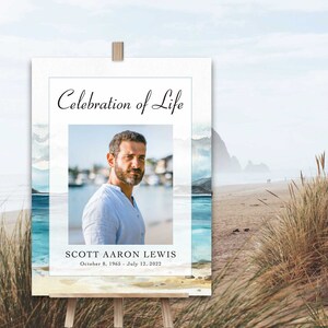 May include: A white sign with a watercolour background of blue and white waves. The sign says "Celebration of Life" and features a photo of a man with a beard. The text below the photo reads "Scott Aaron Lewis, October 8, 1965 - July 12, 2022".