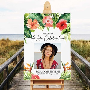 May include: A white sign with a tropical floral border and a photo of a woman in a black hat. The sign reads "Welcome to A Life Celebration Savannah Smythe September 14, 1974 - August 12, 2020".