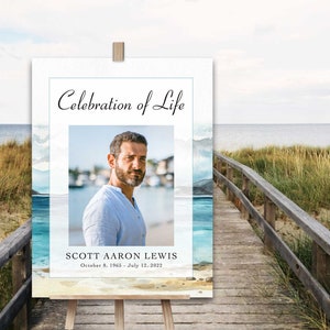 May include: A white sign with a blue watercolour background featuring a beach scene. The sign reads "Celebration of Life" and includes a photo of a man. The text below the photo reads "Scott Aaron Lewis, October 8, 1965 - July 12, 2022".