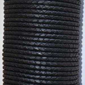 Puede incluir: Un rollo de cord&oacute;n de cuero trenzado negro. El cord&oacute;n est&aacute; enrollado firmemente, creando una forma cil&iacute;ndrica. La textura del cuero es visible, con un patr&oacute;n de hebras entrelazadas. La apariencia general es elegante y oscura.