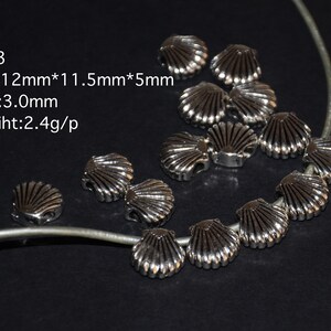 Op de afbeelding: Een close-up van zilverkleurige metalen schelpbedels. De bedels zijn in een cirkel op een zilveren koord gerangschikt. De bedels zijn ongeveer 1,2 cm breed, 1,15 cm lang en 0,5 cm dik. De bedels hebben een gat in het midden met een diameter van 3 mm. De tekst "PA13 Size: 12mm*11.5mm*5mm Hole:3.0mm Wegiht:2.4g/p" is zichtbaar in de afbeelding.