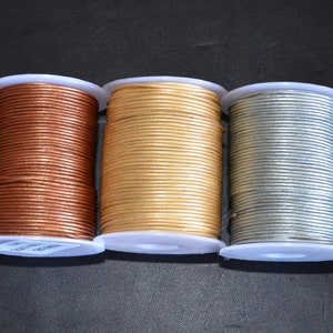 Op de afbeelding: Drie spoelen met lederen koord in verschillende kleuren: koper, goud en zilver. Het koord is om witte kunststof spoelen gewikkeld.
