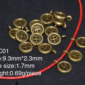 Può includere: Bottoni color ottone antico con quattro fori e un anello per il fissaggio. I bottoni sono esposti su un cordino rosso su sfondo scuro. Il testo "CCC01" e i dettagli delle dimensioni sono visibili.