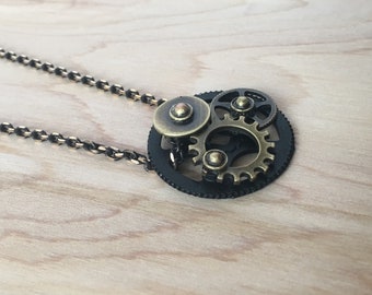 May include: A gold-toned necklace with a pendant featuring a cluster of black and gold gears. The gears are connected and have a steampunk aesthetic.