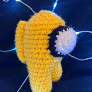 May include: A yellow crocheted plush toy shaped like the character from the video game Among Us. The toy has a black and white eye.