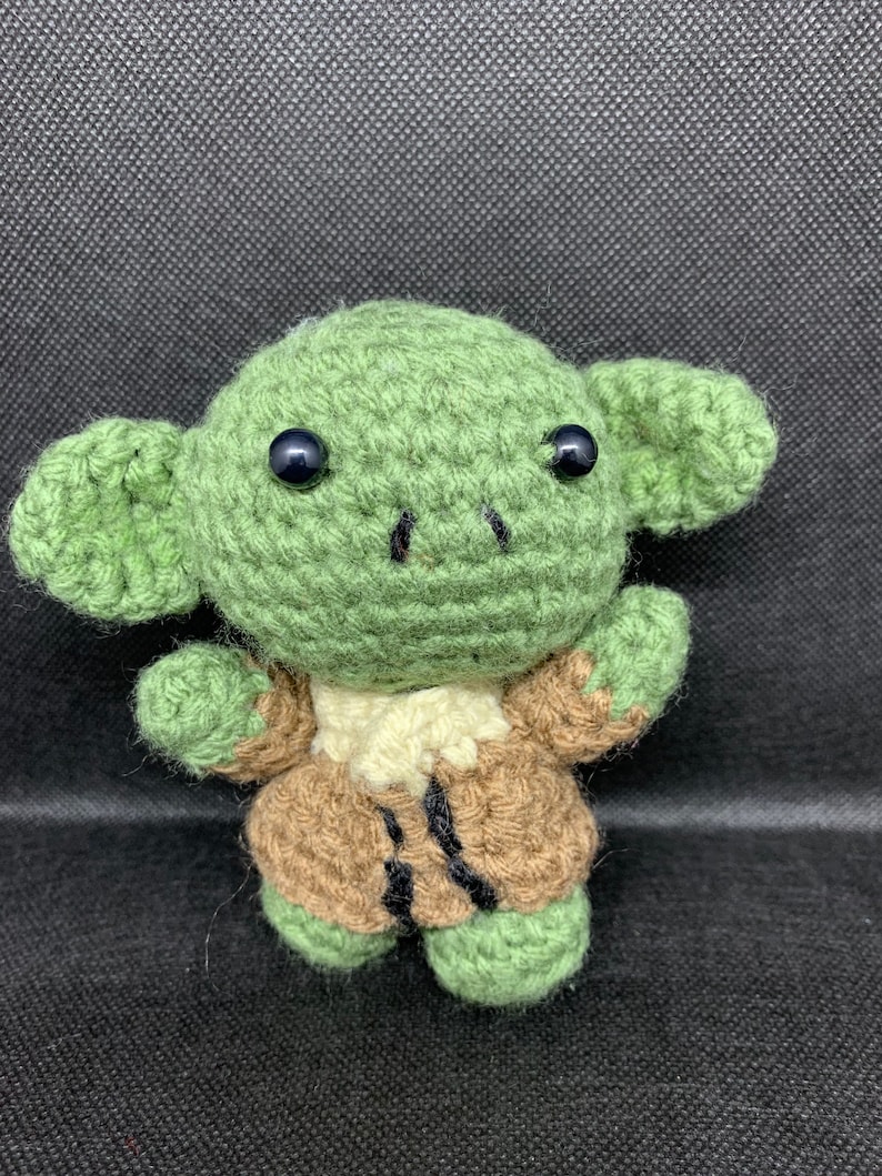 May include: A green crocheted Yoda doll wearing a brown crocheted robe. The doll has black eyes and a small, pointed nose.