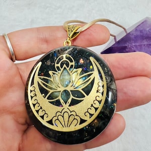 Puede incluir: Un colgante dorado con un diseño de luna creciente y flor de loto, engastado en una resina negra e iridiscente. El colgante está en un cordón beige.