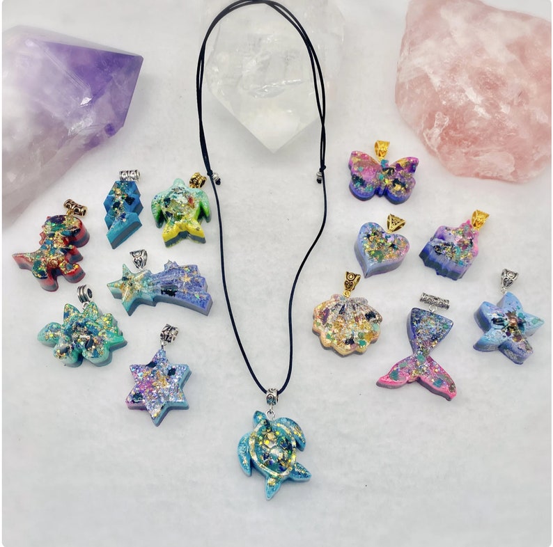 May include: A collection of resin pendants in various shapes including a turtle, dinosaur, butterfly, heart, seashell, mermaid tail, and stars. Each piece features colorful glitter and metallic flakes. One turtle pendant is attached to a black cord necklace.