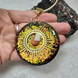 May include: A round black resin pendant featuring a gold-colored sunburst emblem at the center, surrounded by embedded iridescent gold and multicolored foil flakes. The pendant hangs from a light-colored cord with a gold-toned metal bail.