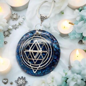 May include: A circular pendant featuring a deep blue, glittery resin base with a silver-toned metal geometric star and circle overlay. The pendant hangs from a silver-toned chain, showcasing a complex, layered design with sharp, angular lines and concentric circles.